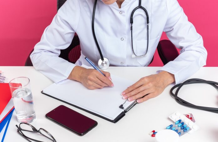 pleased adult slavic female doctor in medical robe with stethoscope sitting at desk with office tools writing on clipboard isolated on pink background with copy space